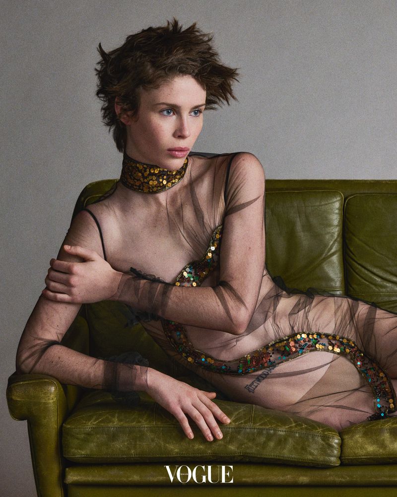 Woman in sheer embellished dress lying on a green vintage couch in a fashion editorial portrait. Studio lighting and neutral background.