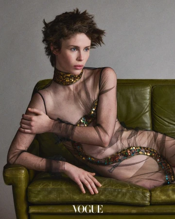 Woman in sheer embellished dress lying on a green vintage couch in a fashion editorial portrait. Studio lighting and neutral background.