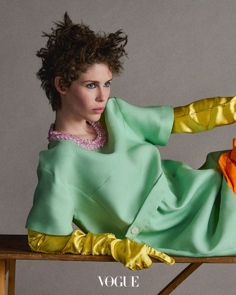 Portrait of a stylish adult female with short hair wearing a mint green dress with yellow gloves and a pink beaded necklace.