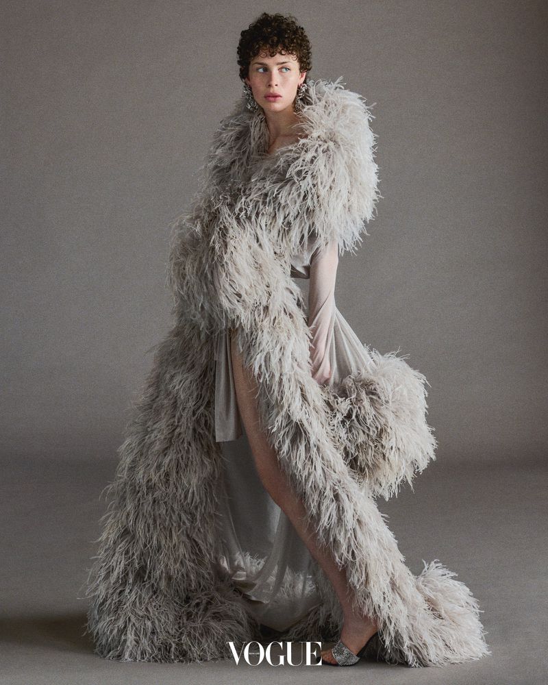 Woman wearing an extravagant, feathered light-grey coat and dress with short curly hair in a studio environment with a neutral background.