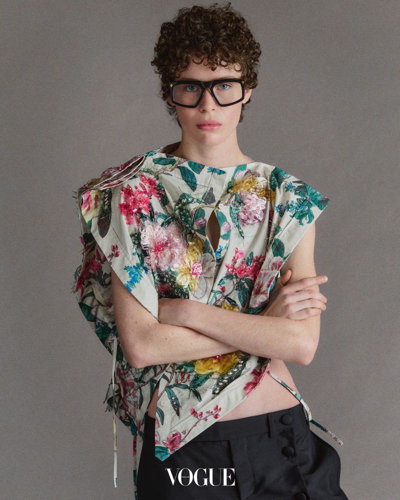 Person with curly hair and glasses wearing a floral top with dark pants in a studio environment, featuring the 'Vogue' logo.