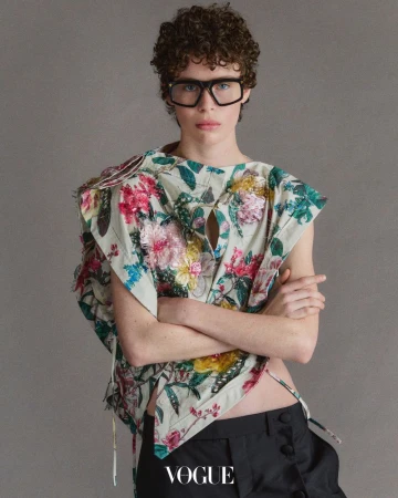 Person with curly hair and glasses wearing a floral top with dark pants in a studio environment, featuring the 'Vogue' logo.