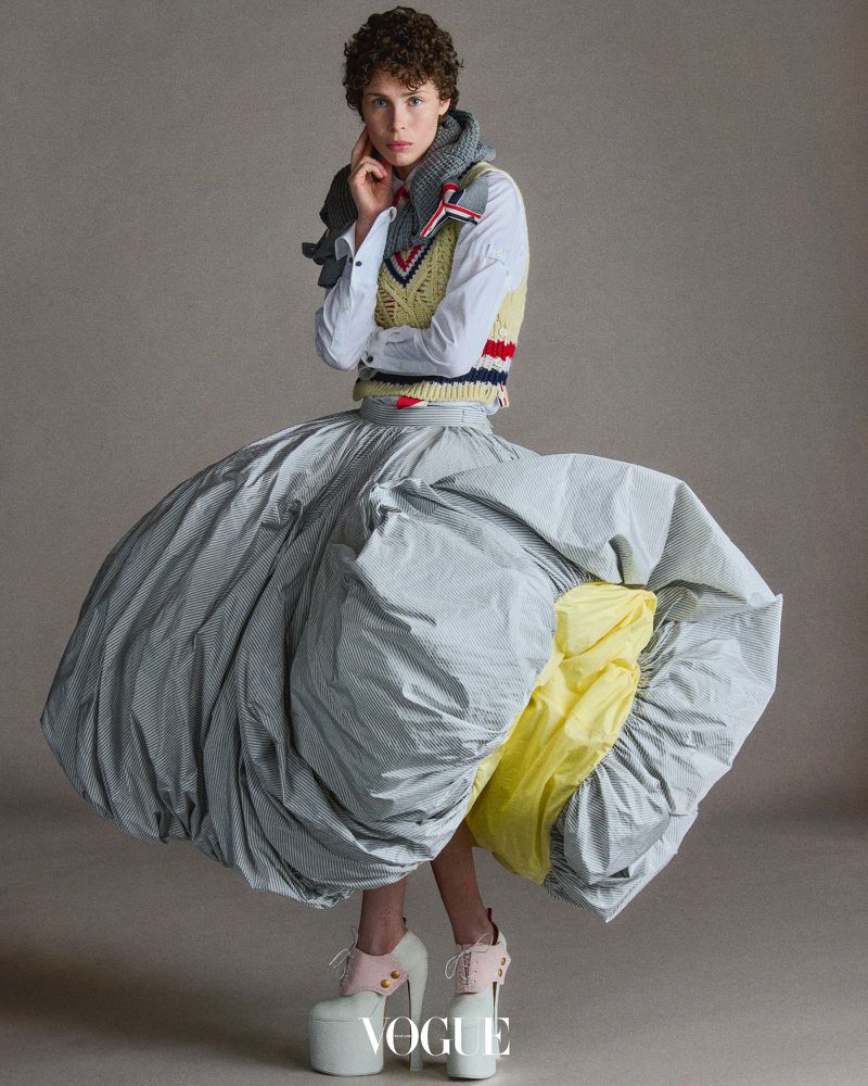 Fashion editorial shot of a model posing in an exaggerated striped balloon skirt, sweater vest, collared shirt, and platform heels in a studio.