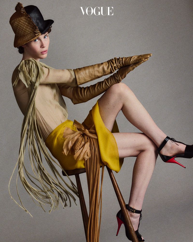 A fair-skinned woman poses in avant-garde fashion including hat, gloves, dress, and heels against a grey backdrop for Vogue magazine.