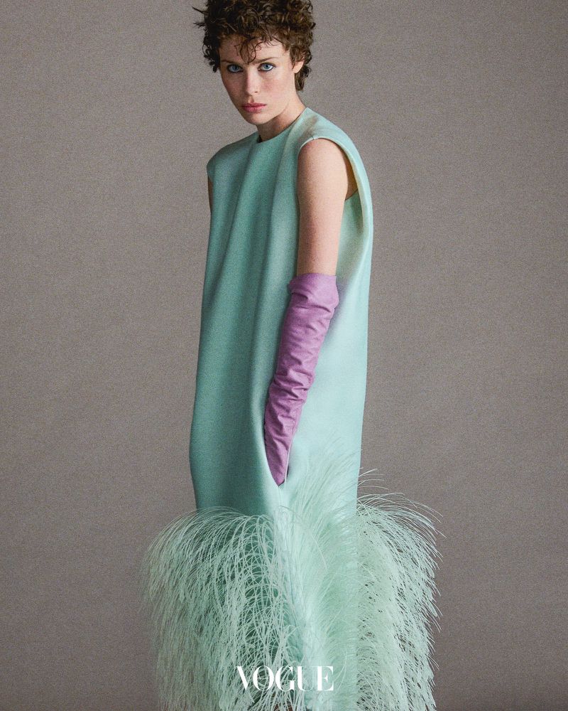 A woman poses in a light blue dress with feather trim and lilac gloves in a studio, creating a modern, elegant fashion image.