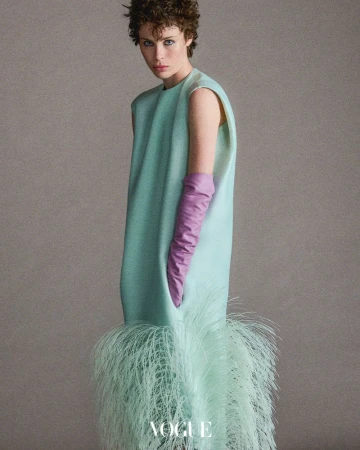 A woman poses in a light blue dress with feather trim and lilac gloves in a studio, creating a modern, elegant fashion image.