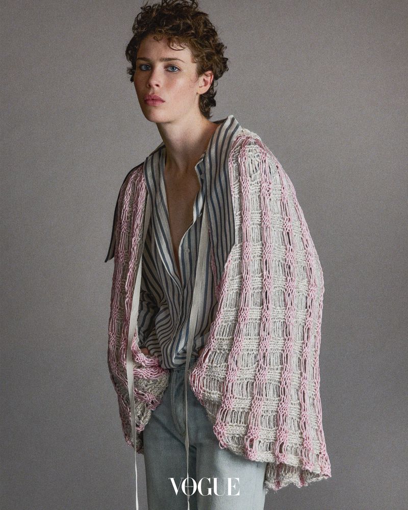 A young person in a striped shirt and knit cardigan poses against a grey backdrop in a fashion editorial shot with soft lighting.