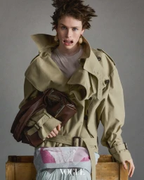 Model wearing a structured beige trench coat and holding a brown leather bag against a neutral grey backdrop