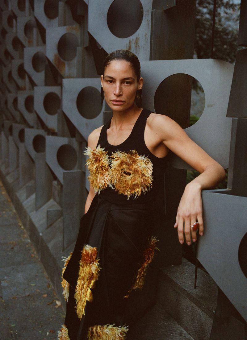 Elegant woman in a black dress with feathered embellishments poses against a geometric metal wall in natural light.
