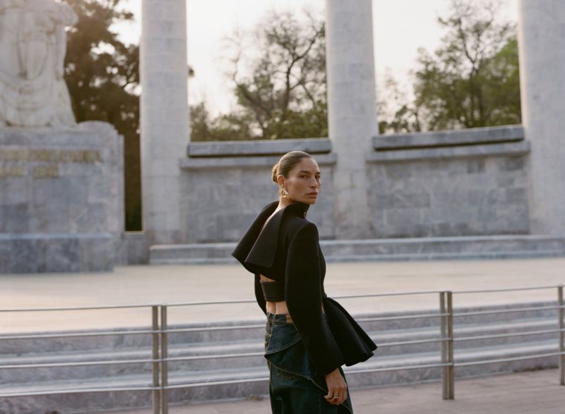 Adult female in architectural outdoor setting. Woman in cropped top and jeans looking over shoulder with neutral expression in daylight.