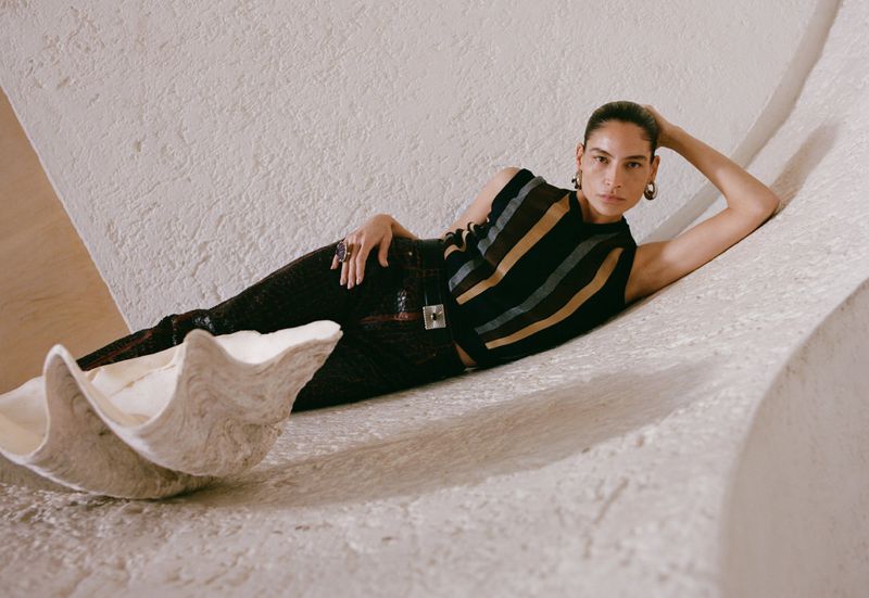 Adult female fashion model in a striped top and black pants lying on a white sculpted surface near a large clam shell indoors.