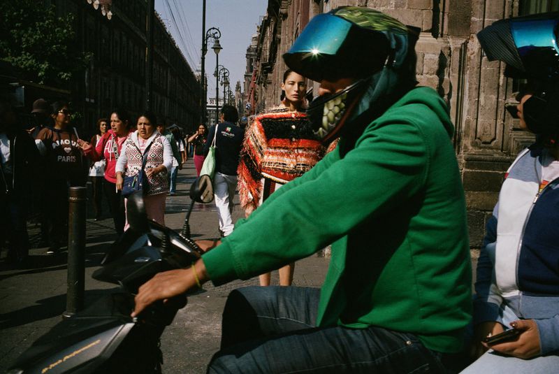 City street scene featuring a person on a moped with a helmet, other pedestrians in the background, and aged buildings on the side.