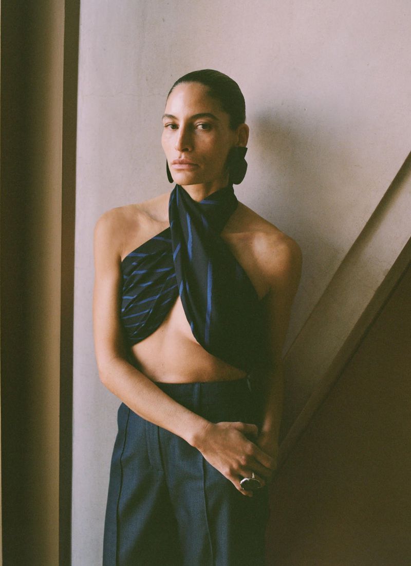 Fashionable woman wearing a blue striped halter top and navy pants poses in front of a textured wall with neutral and warm colors.