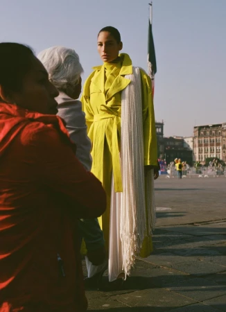 Woman in a yellow coat poses in an urban square, featuring a Mexican flag, buildings, and people in the background on a sunny day.