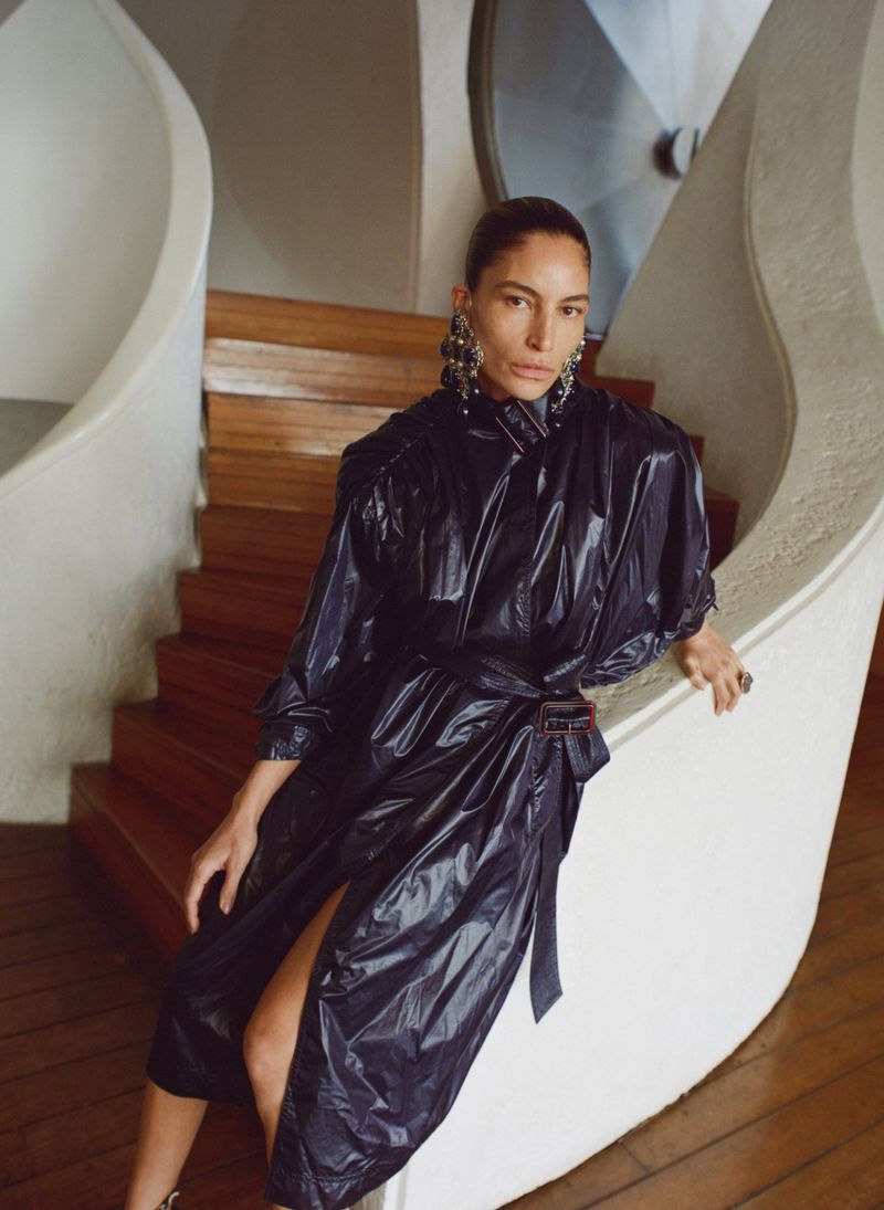 A woman in a blue, shiny coat poses on a staircase, featuring large earrings and a ring, bathed in soft, natural light.