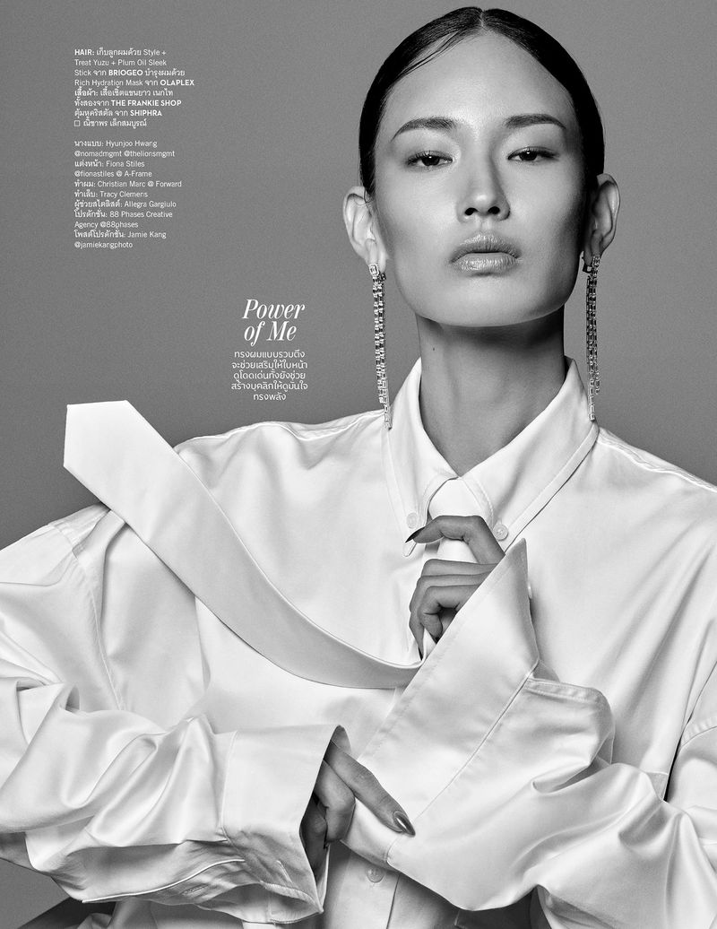 Black-and-white portrait of an Asian woman with long earrings wearing a white oversized shirt against a grey background.