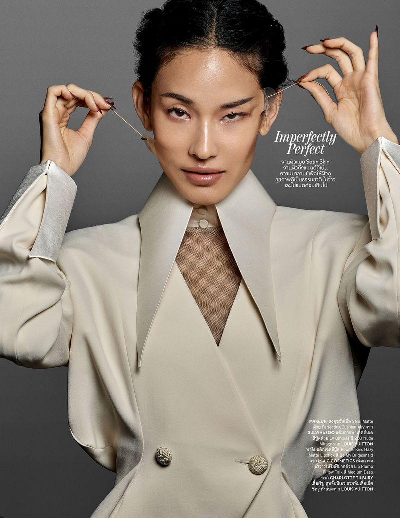 Portrait of an adult woman wearing a white suit jacket with a sheer shirt, applying tape to her face in a neutral studio setting.