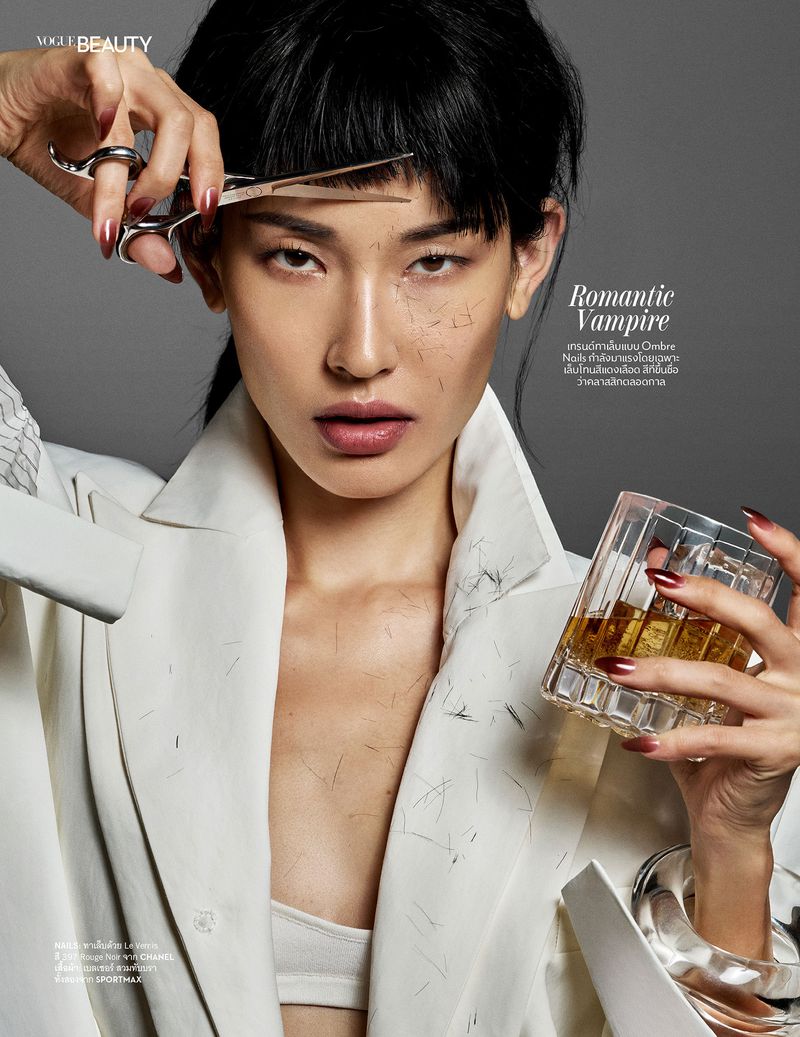 Vogue Beauty portrait of Asian woman cutting bangs with scissors, holding glass of alcohol, against a grey background in studio lighting.
