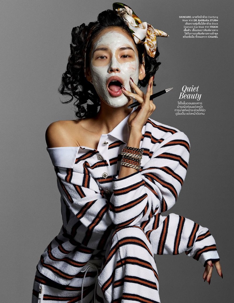 Portrait of woman with face mask and a scarf, wearing a brown and white striped top and holding an makeup brush in her hand.