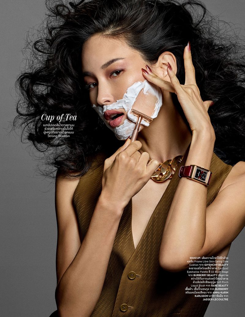 Glamorous woman shaves face with vintage razor, sporting a gold chain and vest in a beauty editorial on a gray backdrop.