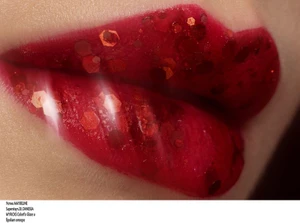 Close-up macro shot of glossy red lips covered with chunky hexagonal red glitter flakes for an editorial beauty look.