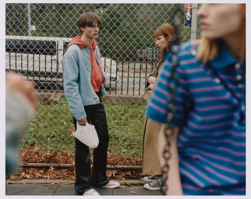 A group of three fashionably dressed people in an outdoor urban setting, one holding a white plastic bag, standing on a sidewalk.