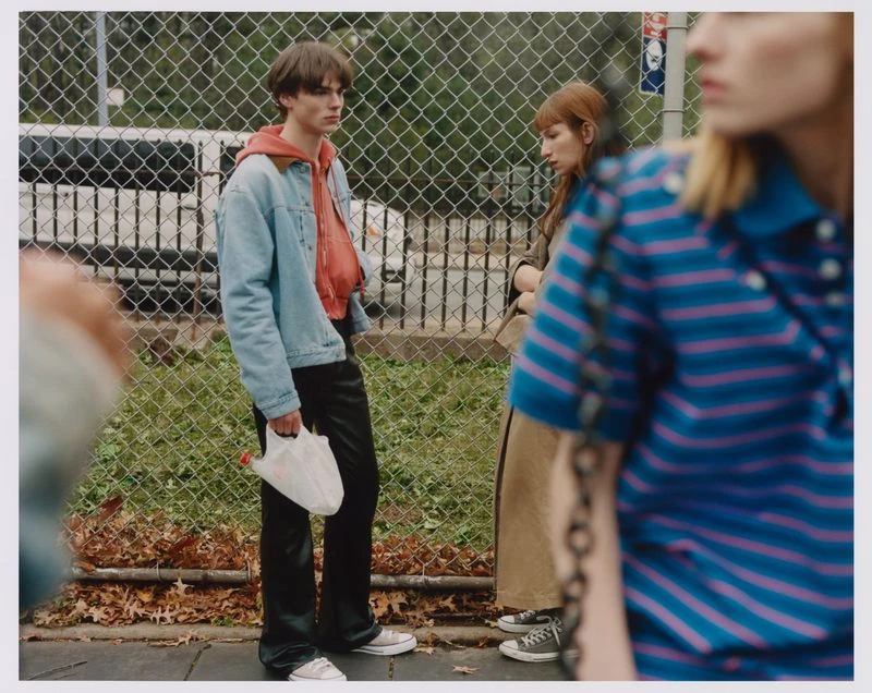 A group of three fashionably dressed people in an outdoor urban setting, one holding a white plastic bag, standing on a sidewalk.