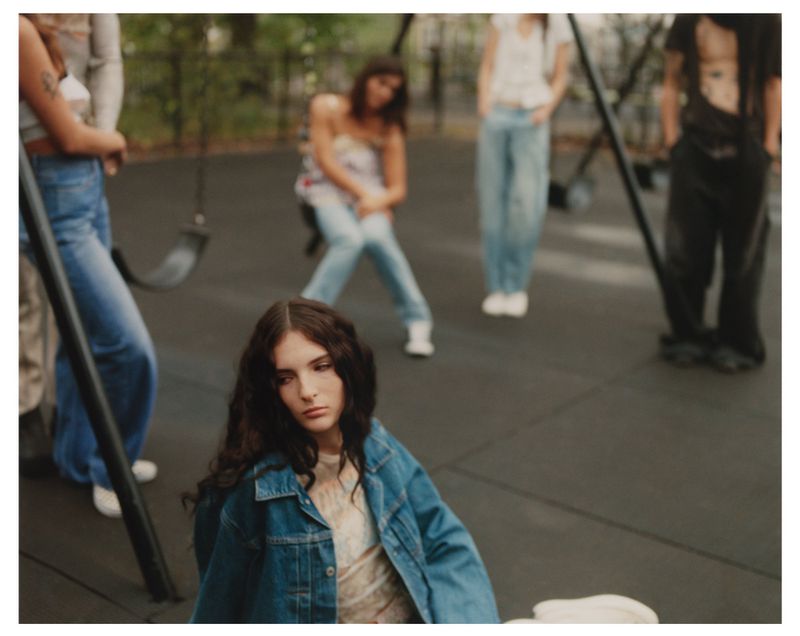 A group of young adults gathers in an outdoor park setting, one female with long hair and a denim jacket is seated in the foreground.