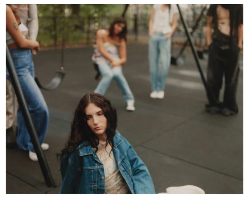 A group of young adults gathers in an outdoor park setting, one female with long hair and a denim jacket is seated in the foreground.