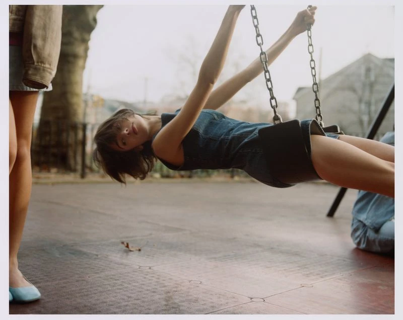 Woman on a swing in a playground wearing a denim dress, with another person standing nearby. The scene is captured at eye-level outdoors.