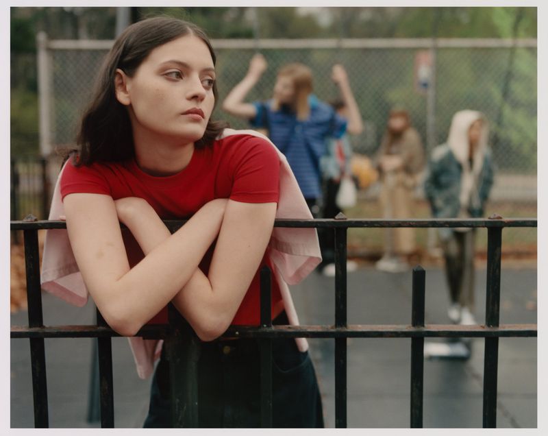 Teen girl in red shirt leaning on black fence in park, with blurred group of people in background, outdoor portrait.