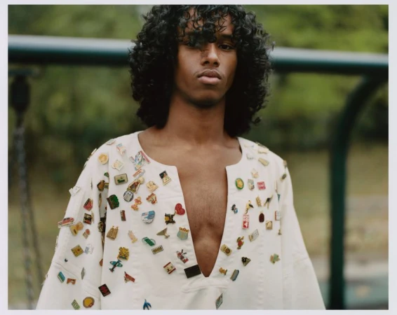 Portrait of a curly-haired person in a white shirt with various pins, posing outdoors against a blurred background of green foliage and playground equipment.