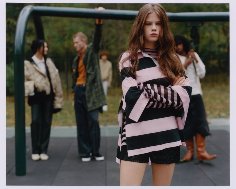 Fashion editorial shot outdoors featuring a young woman in a striped top, with several other people in the blurred background.
