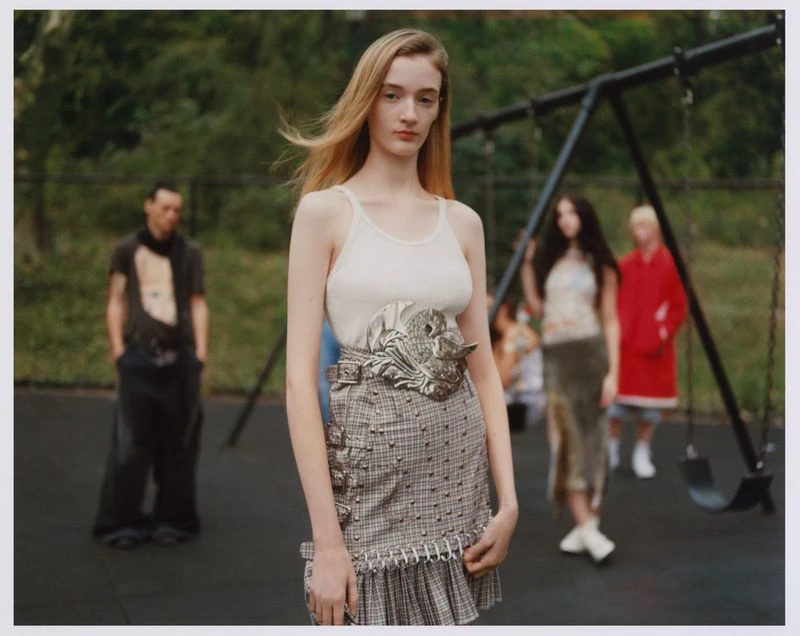 A tall, light-skinned woman with blonde hair poses in a plaid skirt and tank top in front of a blurred playground scene with several people.