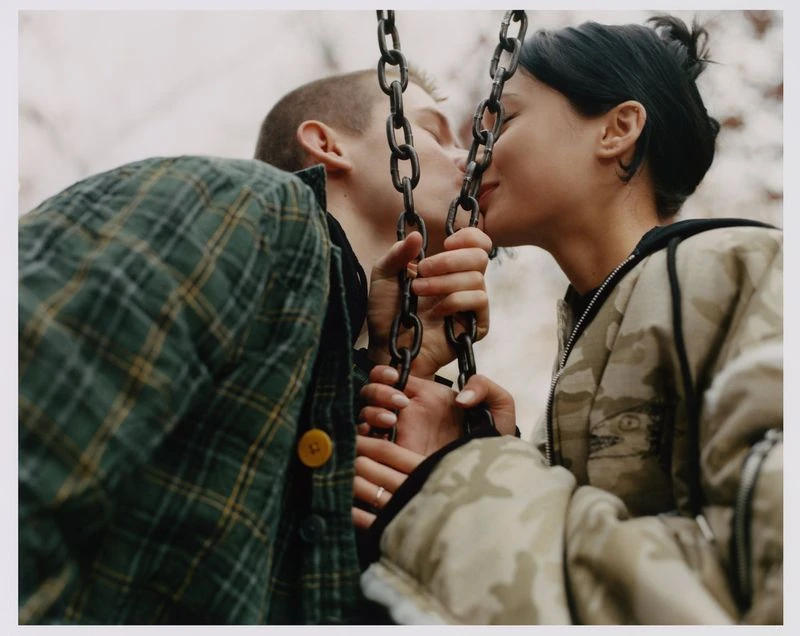 A couple kisses on a swing set, dressed in casual jackets, capturing a candid moment in an outdoor setting with soft, natural lighting.