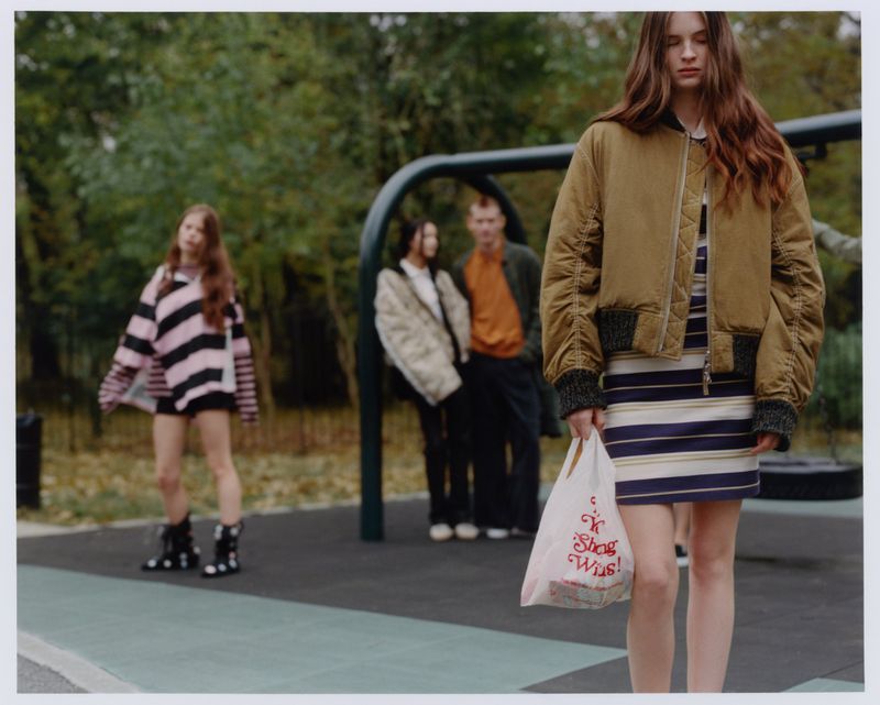 A group of four people in an outdoor setting, with a young woman in the foreground holding a shopping bag and wearing a bomber jacket.