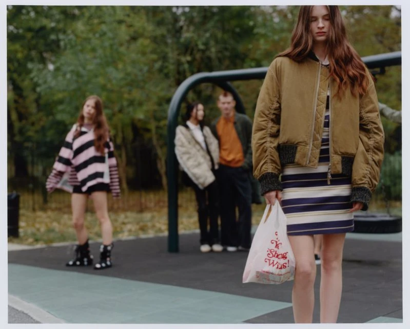 A group of four people in an outdoor setting, with a young woman in the foreground holding a shopping bag and wearing a bomber jacket.