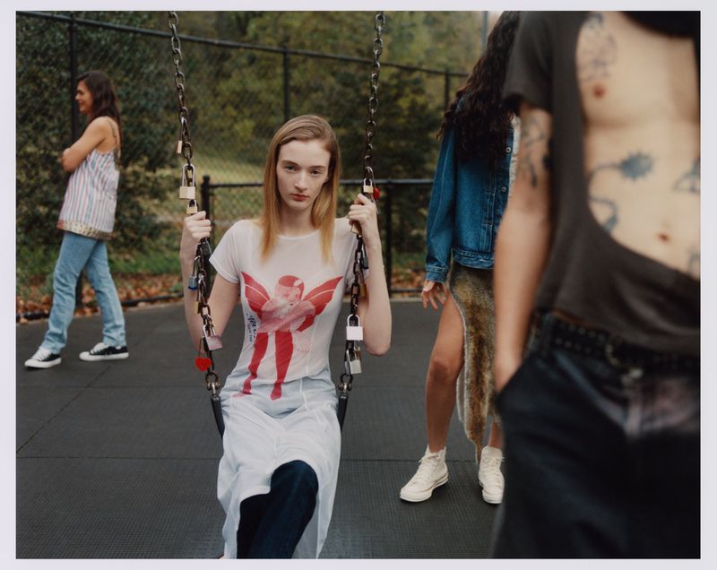 A young woman on a swing with people in the background in an outdoor setting, with fashion-forward clothing and natural lighting.