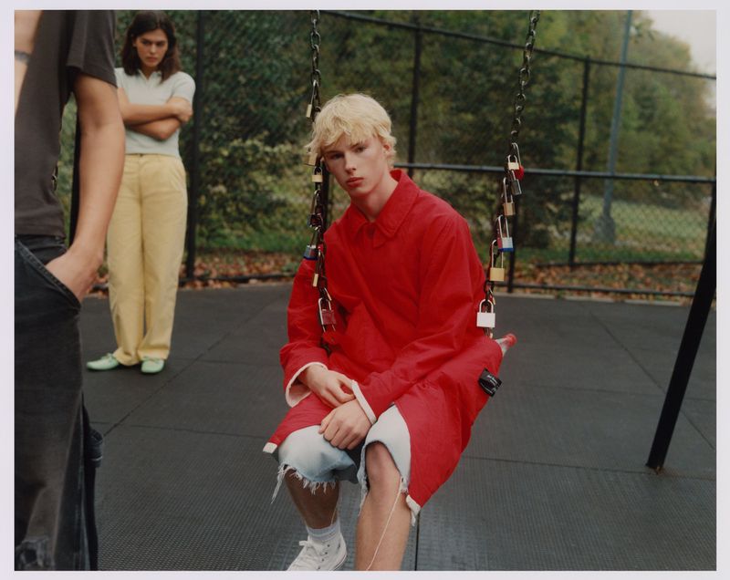 Young person with blonde hair in a red coat sits on a swing with locks attached to the chains in an outdoor park setting with other people.