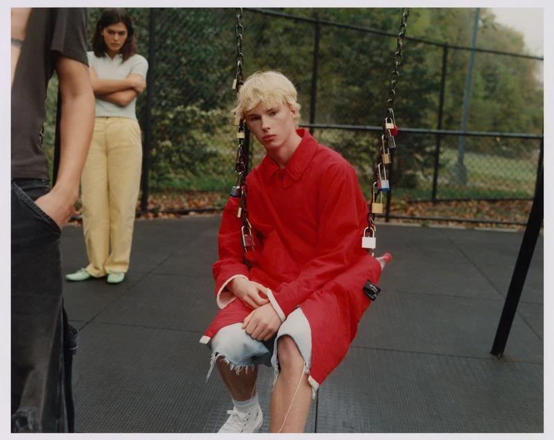 Young person with blonde hair in a red coat sits on a swing with locks attached to the chains in an outdoor park setting with other people.