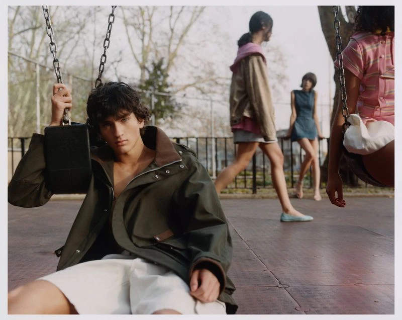 Fashion photo of a young man on a swing in a park with other models in the background, under natural light, styled with an olive jacket.