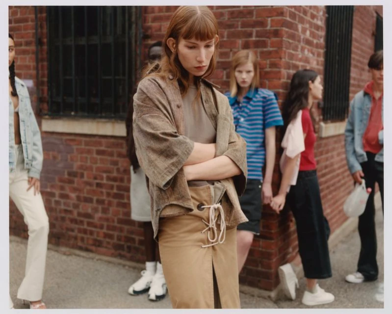 A diverse group of people stands against a brick wall in an urban setting, fashionably dressed in neutral tones and contemporary styles.