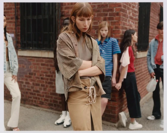 A diverse group of people stands against a brick wall in an urban setting, fashionably dressed in neutral tones and contemporary styles.