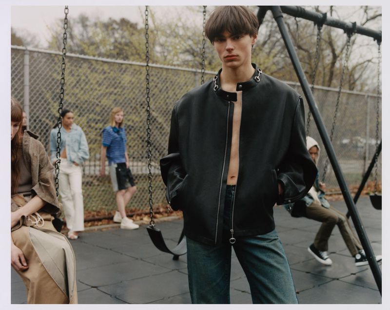 Stylish male model poses in an unzipped black jacket and jeans, captured in a medium shot on a playground swing. 