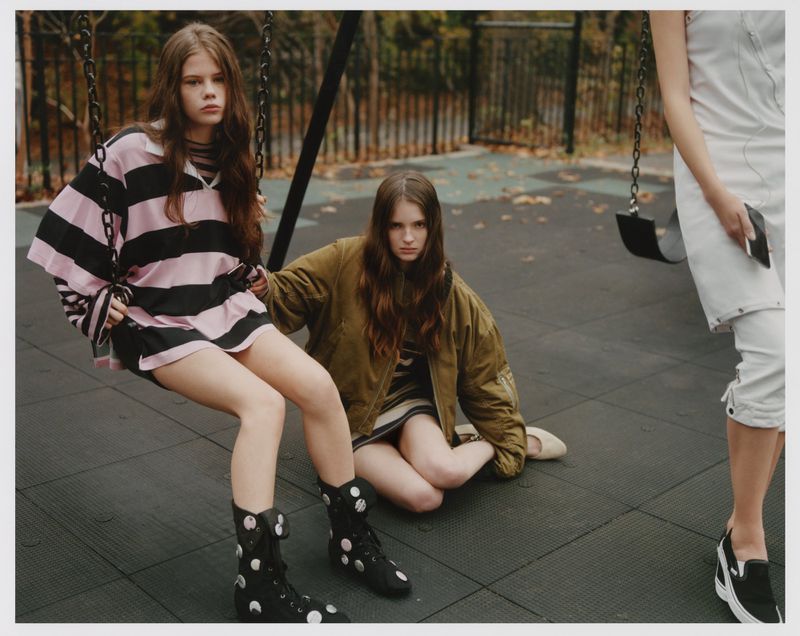 Three young women in a playground setting, with two seated and one standing, wearing casual fashion and posing for a casual shot.