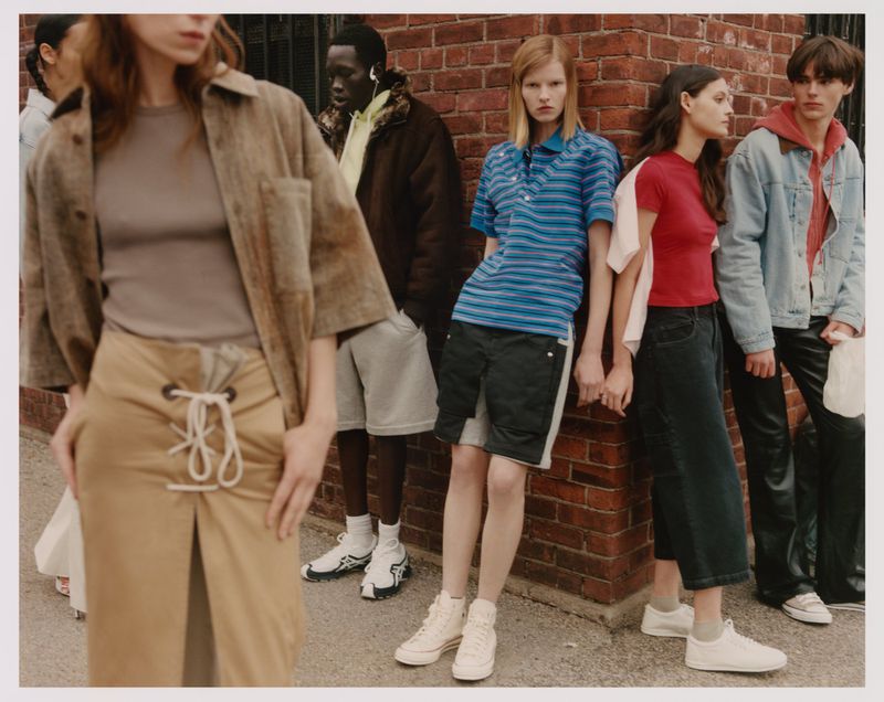 Group of young adult fashion models posing in casual outfits against a red brick wall in an outdoor urban setting with natural lighting.