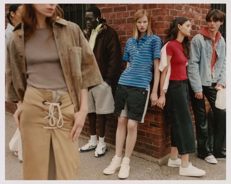 Group of young adult fashion models posing in casual outfits against a red brick wall in an outdoor urban setting with natural lighting.