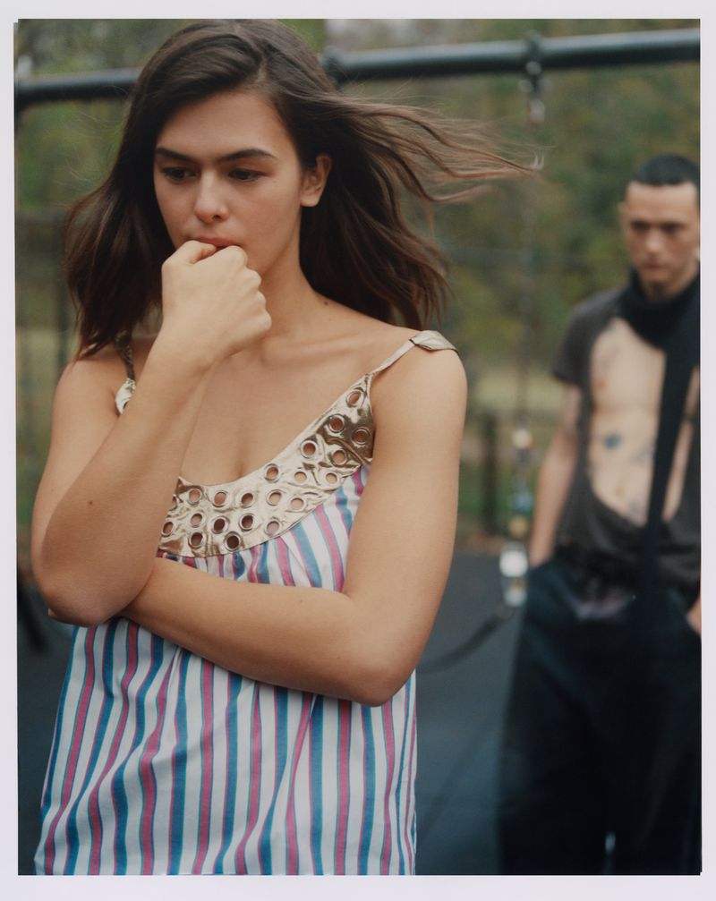 A young woman with brown hair and a striped dress stands outdoors with another person in the background, captured in natural light.