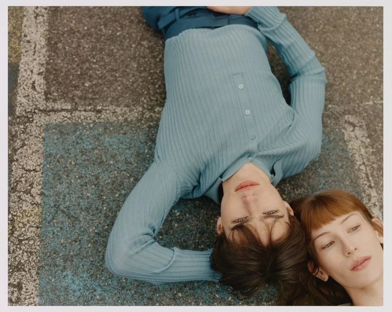 Two people lie on a textured concrete surface, one with dark hair and the other with red hair, wearing matching light blue knitwear.