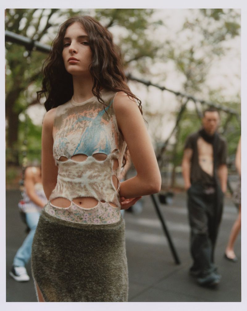 Woman with long brown hair poses in a beige cut-out top and long skirt, with blurred people and playground equipment in the background.